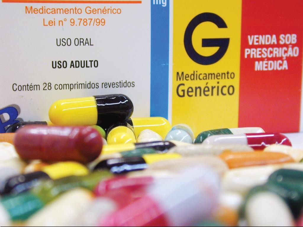 Foto com inúmeras pílulas coloridas espalhadas em frente à uma caixa de medicamento genérico.